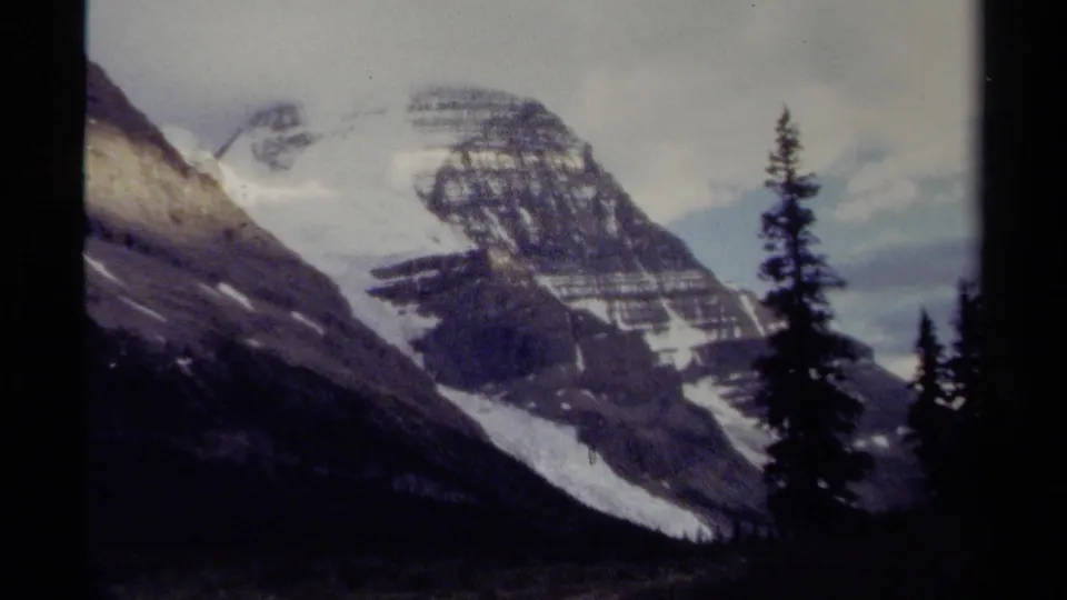 1979 8mm British Columbia mountain view with a snow-streaked rocky peak, dark conifers, and soft overcast light.
