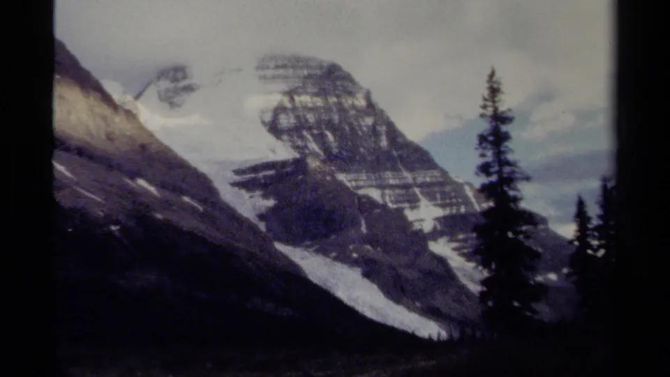 British Columbia 8mm frame showing a layered rocky mountain face with snow patches and conifer trees in front.