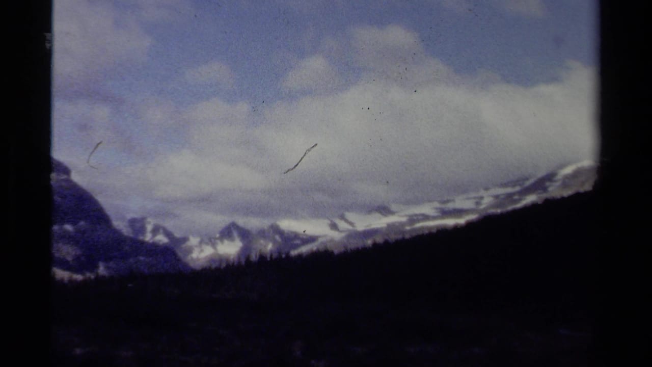 1979 8mm British Columbia mountain vista with snow-covered peaks, blue sky, cloud bands, and dark frame edges.