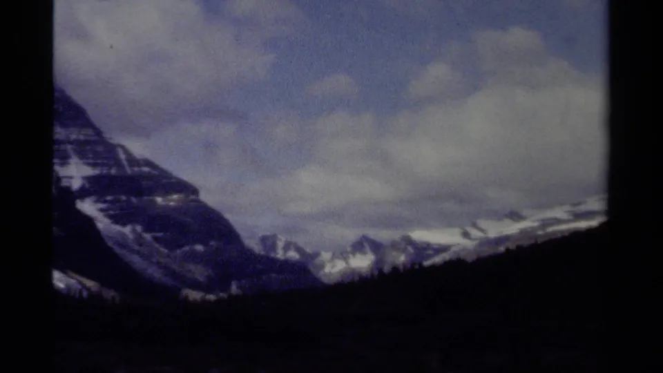 British Columbia mountain view on 8mm film, with a dark foreground slope, distant snow peaks, and soft cloud cover.