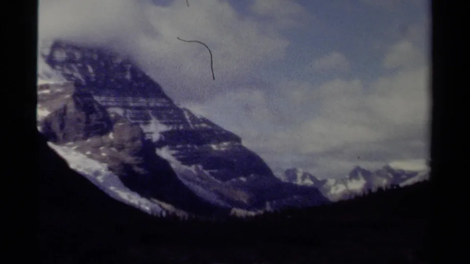 1979 British Columbia 8mm frame showing a broad rocky mountain face with snow patches, distant peaks, and clouded sky.