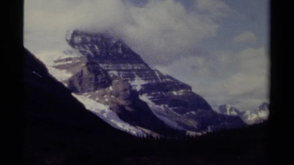 8mm home-movie view of a British Columbia mountain ridge, showing snow streaks, layered rock, clouds, and dark foreground terrain.