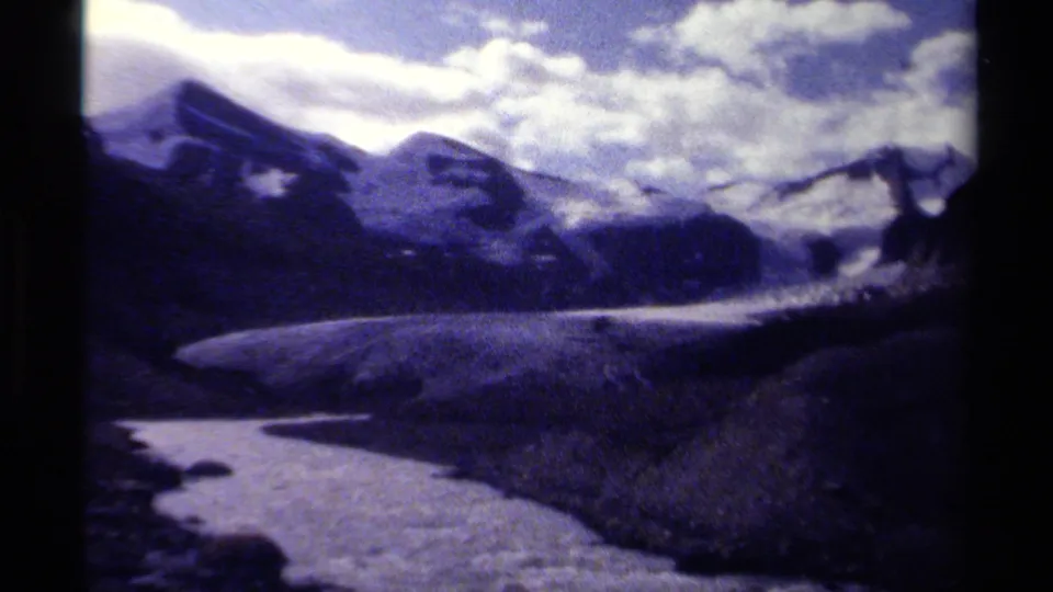 1979 British Columbia 8mm frame showing a wide glacial valley with dark mountain ridges, snow patches, and a bright cloud layer.