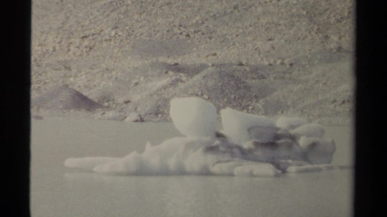 Small iceberg floating near a rocky shoreline on calm water.