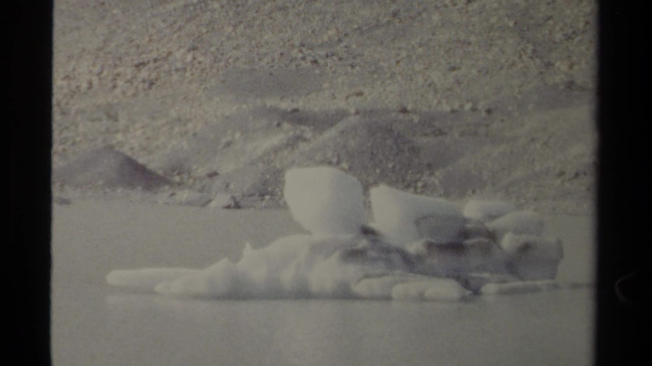 Floating ice mass on water with a muted rocky bank in the background.
