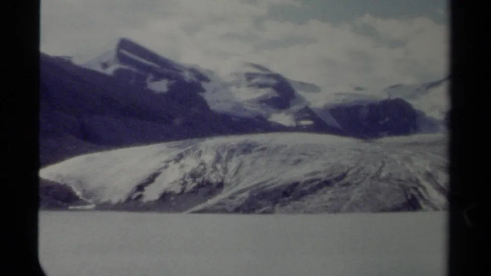 8mm home-movie frame of a glacier and snow-striped mountains across water in British Columbia, with cloudy sky overhead.