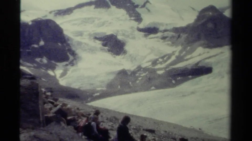 1979 British Columbia home-movie frame with distant visitors seated on rocks below a broad snowy glacier landscape and rugged peaks.
