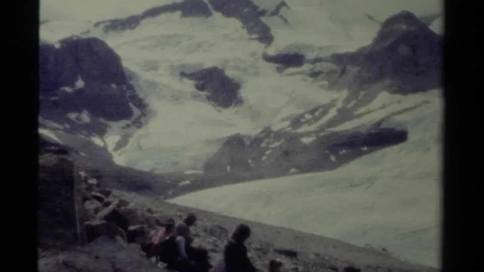 Faded 8mm British Columbia vacation frame showing a wide snowy alpine view with distant people seated on a rocky slope.