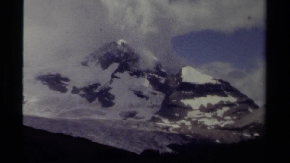 1979 British Columbia 8mm frame showing distant snow-covered mountains beneath soft clouds and a cool blue-gray cast.