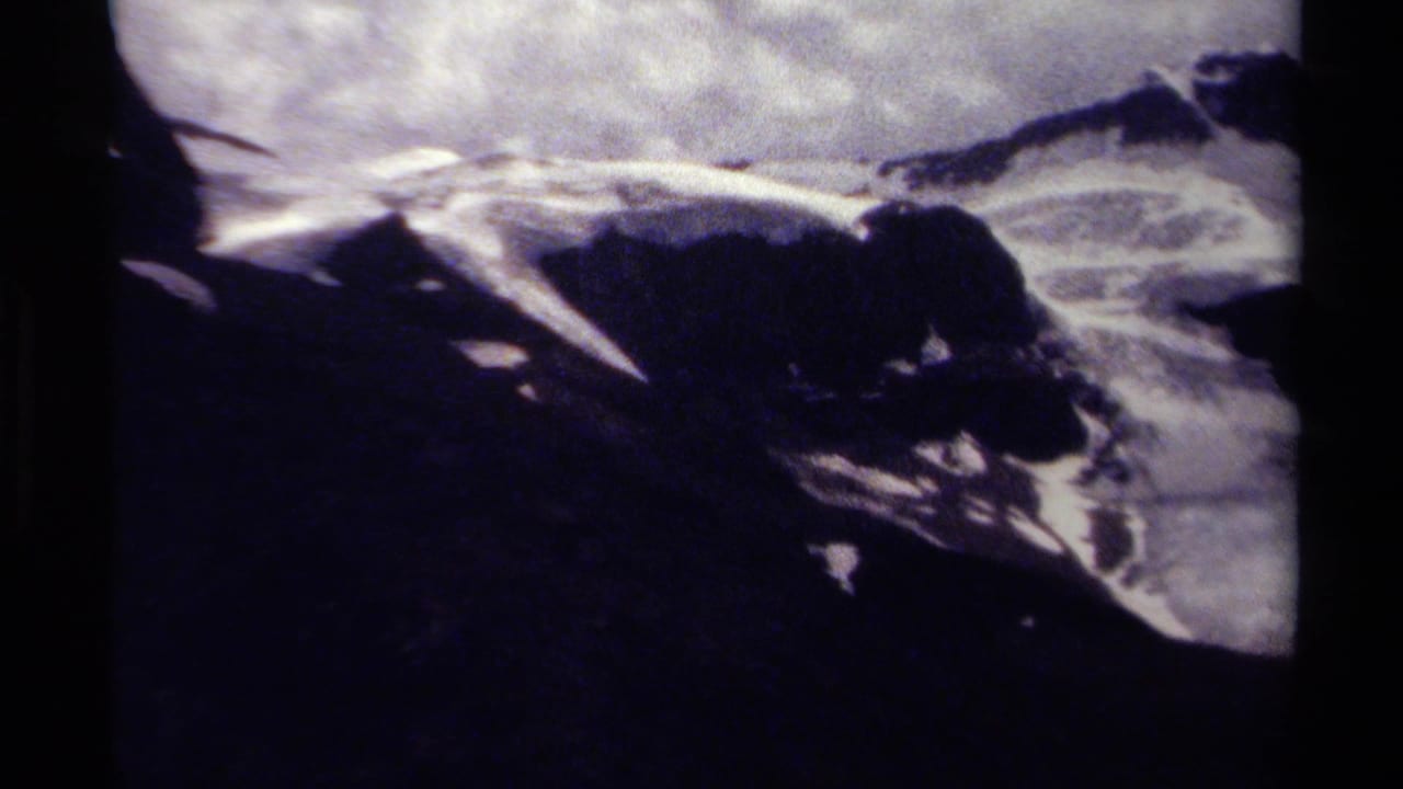 Grainy 8mm frame of British Columbia mountain terrain, showing snowfields, dark rock, and overcast light.