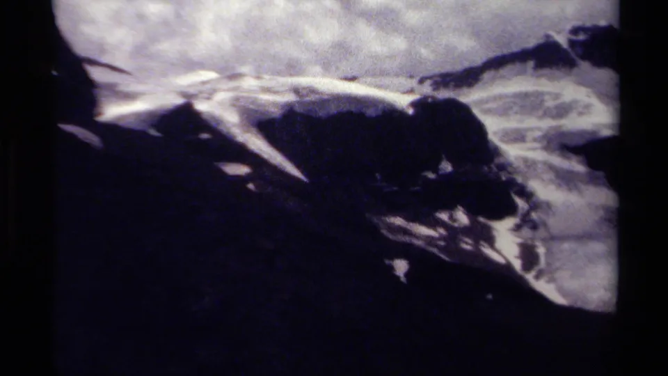 Grainy 8mm frame of British Columbia mountain terrain, showing snowfields, dark rock, and overcast light.