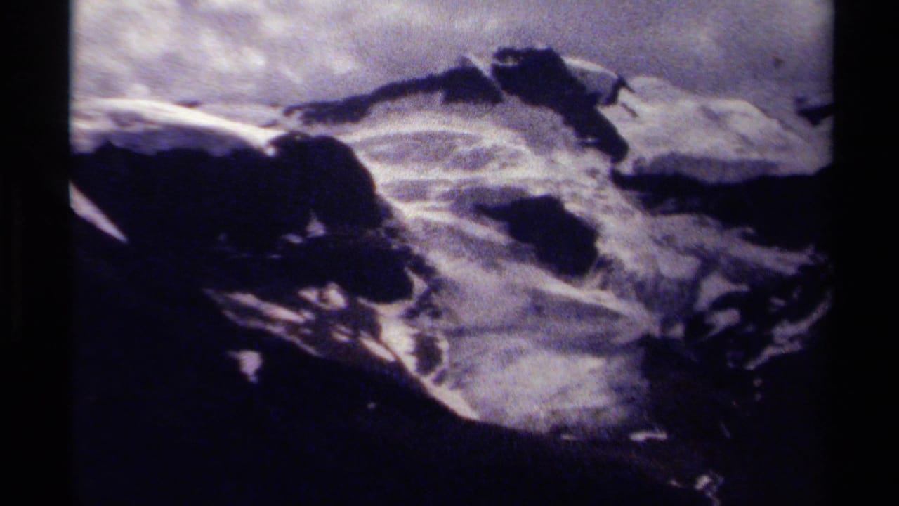 1979 8mm home-movie frame of a rugged British Columbia mountain ridge with snow patches, clouds, grain, and dark film edges.