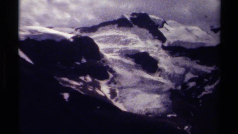 1979 8mm home-movie frame of a rugged British Columbia mountain ridge with snow patches, clouds, grain, and dark film edges.