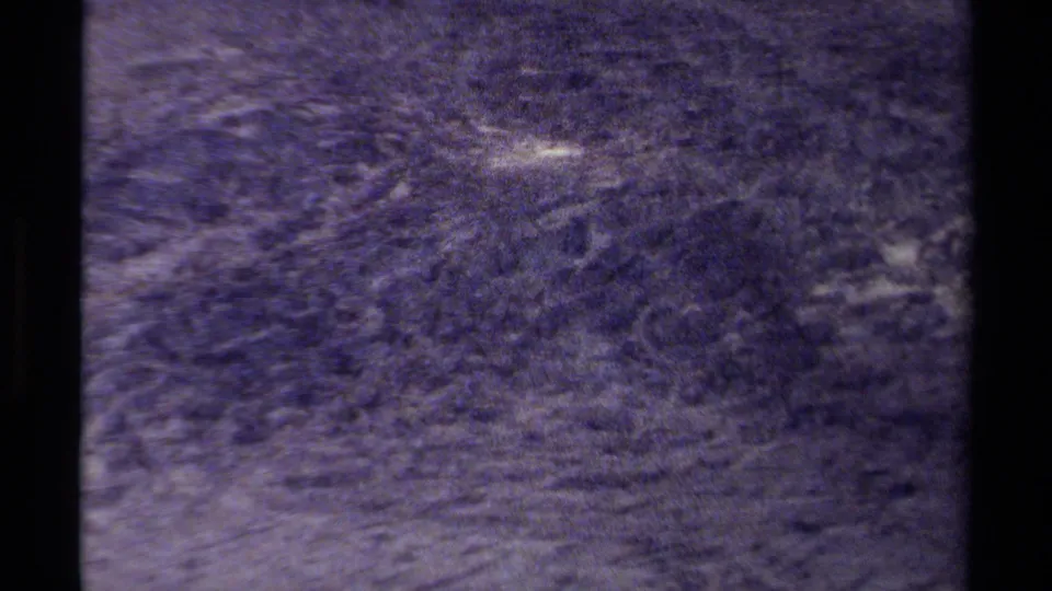 1979 8mm frame showing a blurred British Columbia alpine surface with faint slope texture, a pale snow patch, film grain, and faded color.