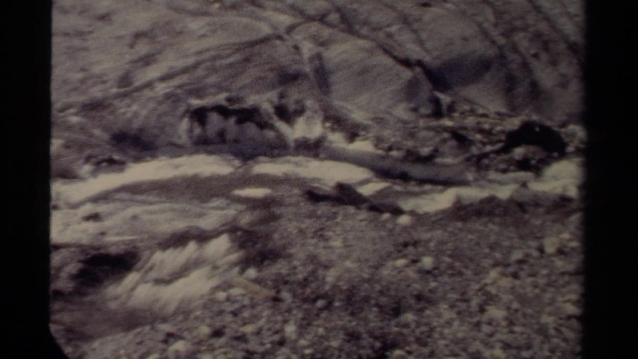 Fast water crossing a rocky stream bed with sloped banks behind it.