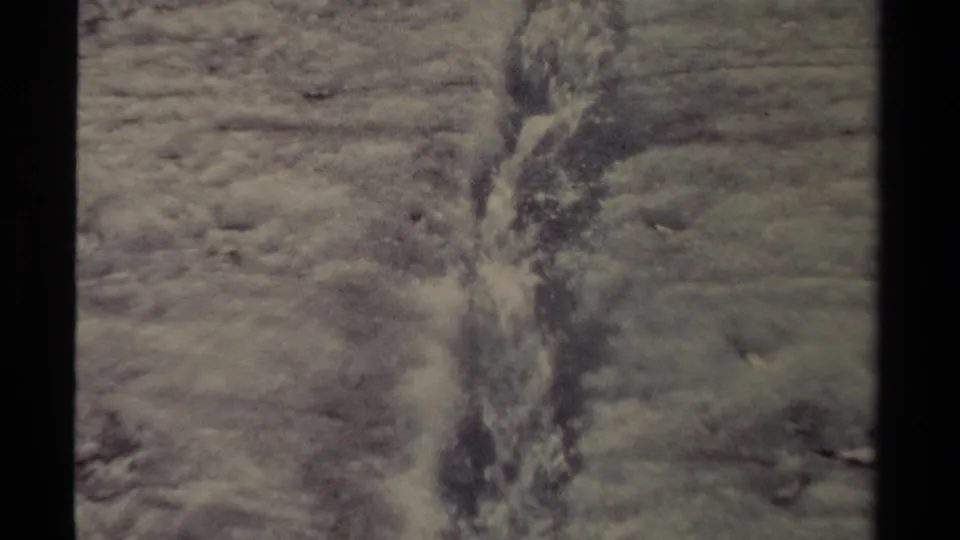 Home-movie frame from British Columbia showing meltwater moving through a narrow icy groove on frozen terrain.