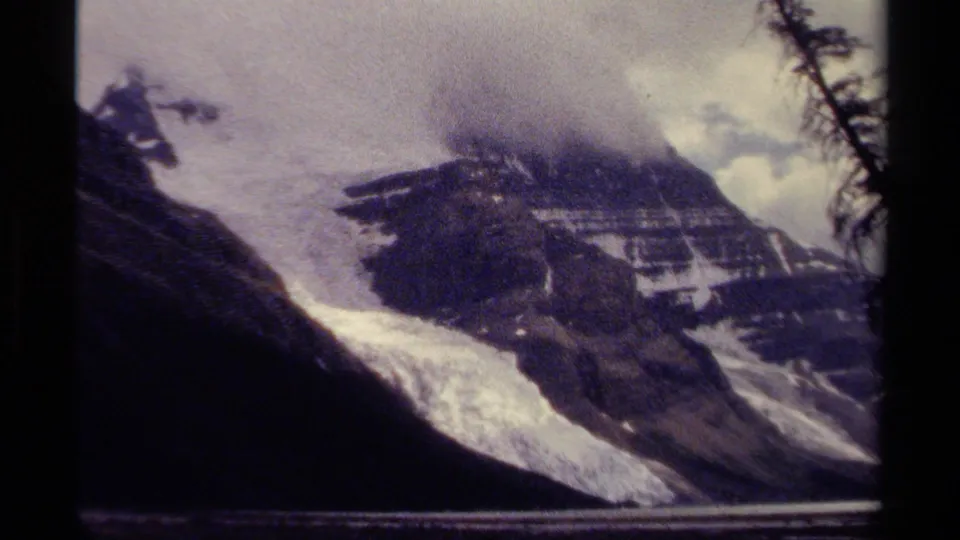 1979 British Columbia mountain view with a bright snowfield, dark ridge, low cloud, and a tree branch at the frame edge.