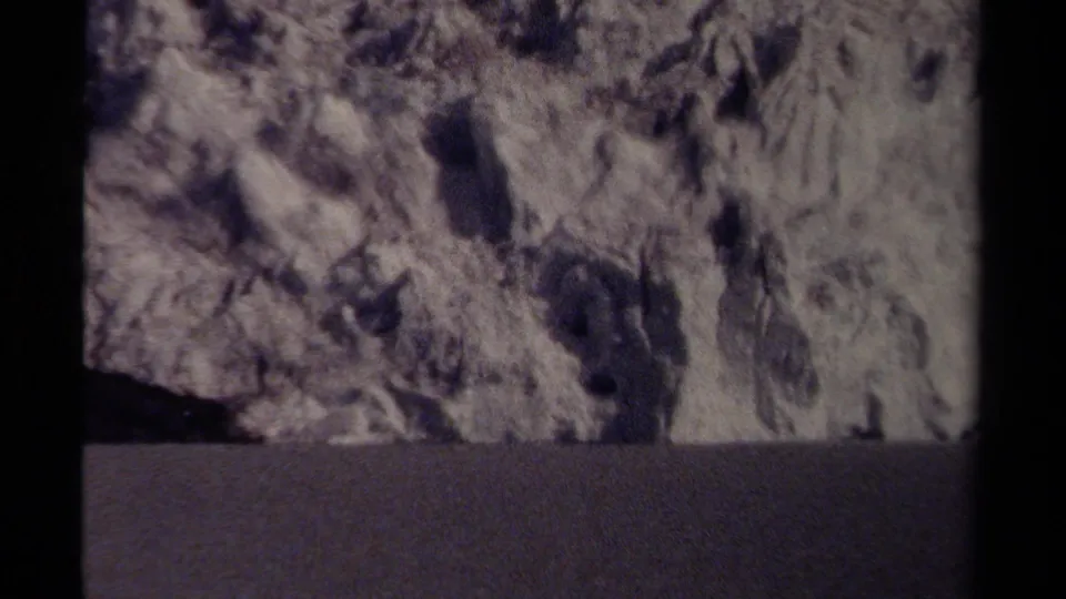 Grainy 8mm frame of a large, rugged rock formation near a road in British Columbia, with the composition held steady.