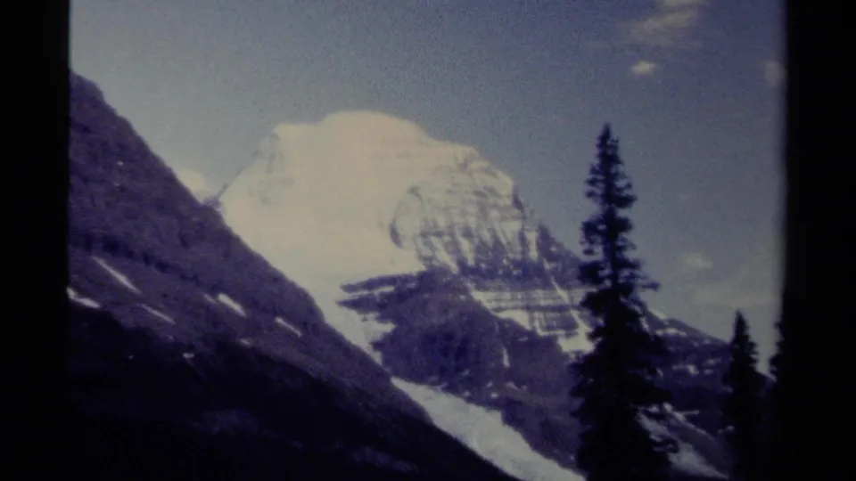 1979 8mm British Columbia mountain view with a snow-capped peak, dark rocky slope, evergreen silhouettes, and pale blue sky.