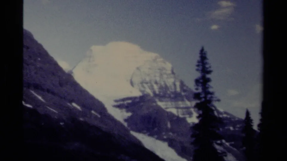 Vintage 8mm view of a British Columbia alpine landscape, showing a bright snow-covered peak beyond dark slopes and evergreen tree silhouettes.