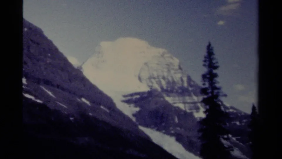 1979 8mm British Columbia mountain view with a pale snowy peak, dark alpine slopes, and evergreen silhouettes in the foreground.