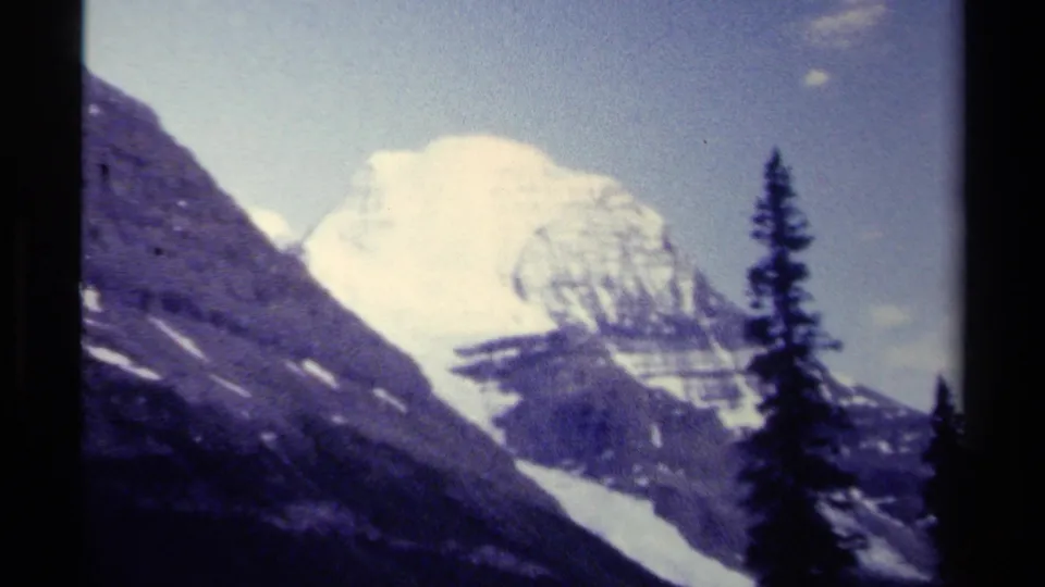 1979 British Columbia home-movie frame showing a soft snowy mountain peak beyond dark slopes, trees, and narrow black film borders.