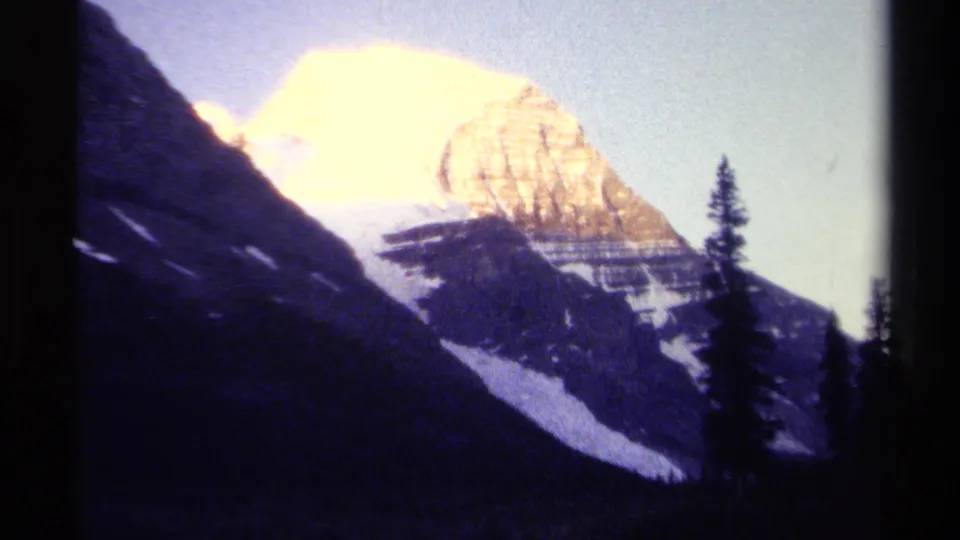 British Columbia 8mm frame from 1979 with golden sunlight on a snow-capped mountain, dark ridges, evergreen silhouettes, and visible film grain.
