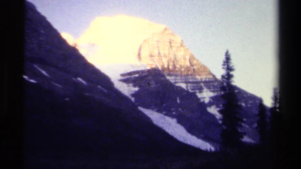 1979 8mm view of a snow-covered mountain peak in British Columbia, framed by dark slopes and evergreen trees.