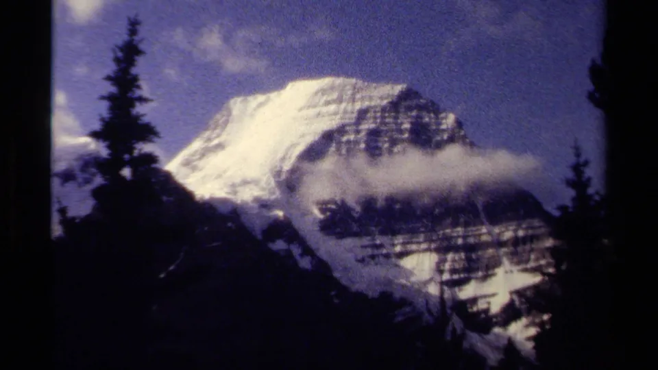 1979 British Columbia 8mm frame showing a snow-covered mountain face, dark pines, and low cloud across the slope.