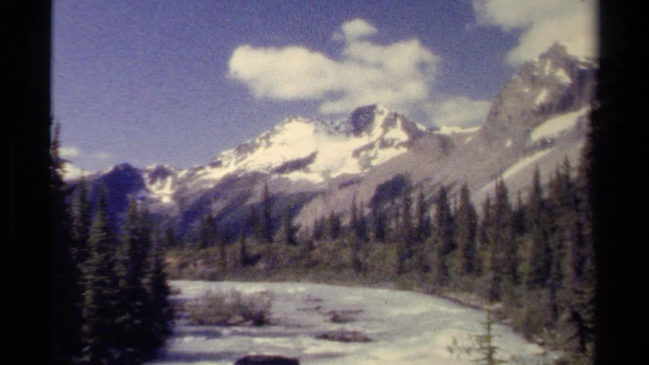 1979 British Columbia 8mm frame with alpine peaks, a curving river, forested banks, and scattered clouds.