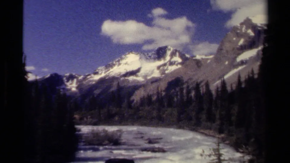 1979 British Columbia 8mm frame showing snow-streaked mountains, evergreen slopes, and a bright river below.