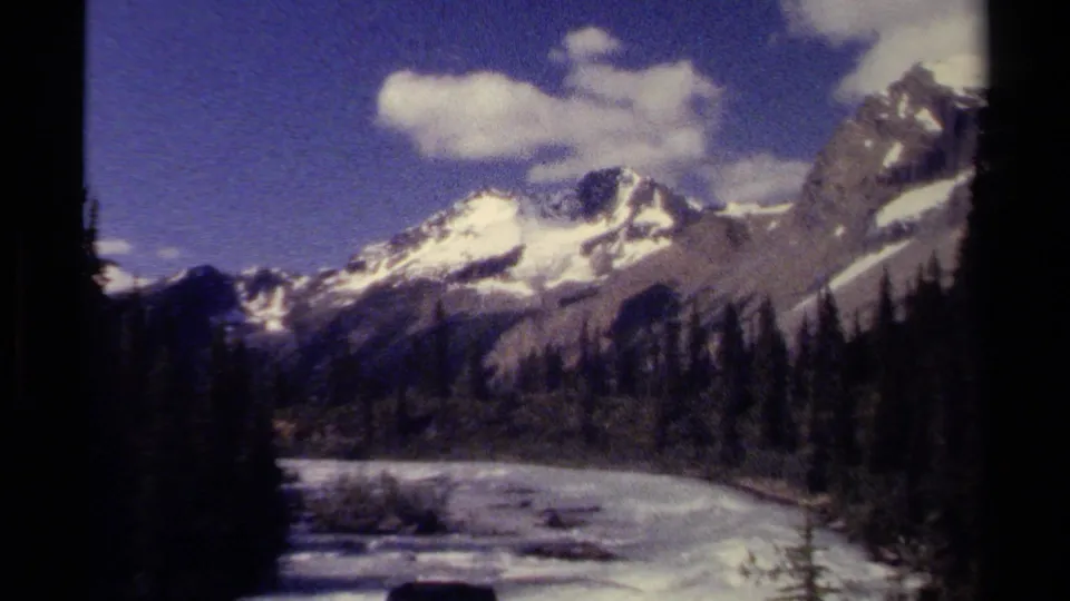 1979 British Columbia 8mm frame of snow-covered alpine peaks above a forested river valley.