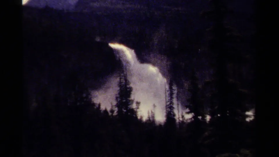 Wider 1979 British Columbia frame showing a waterfall behind evergreen silhouettes, with mist rising against a dark forest background.