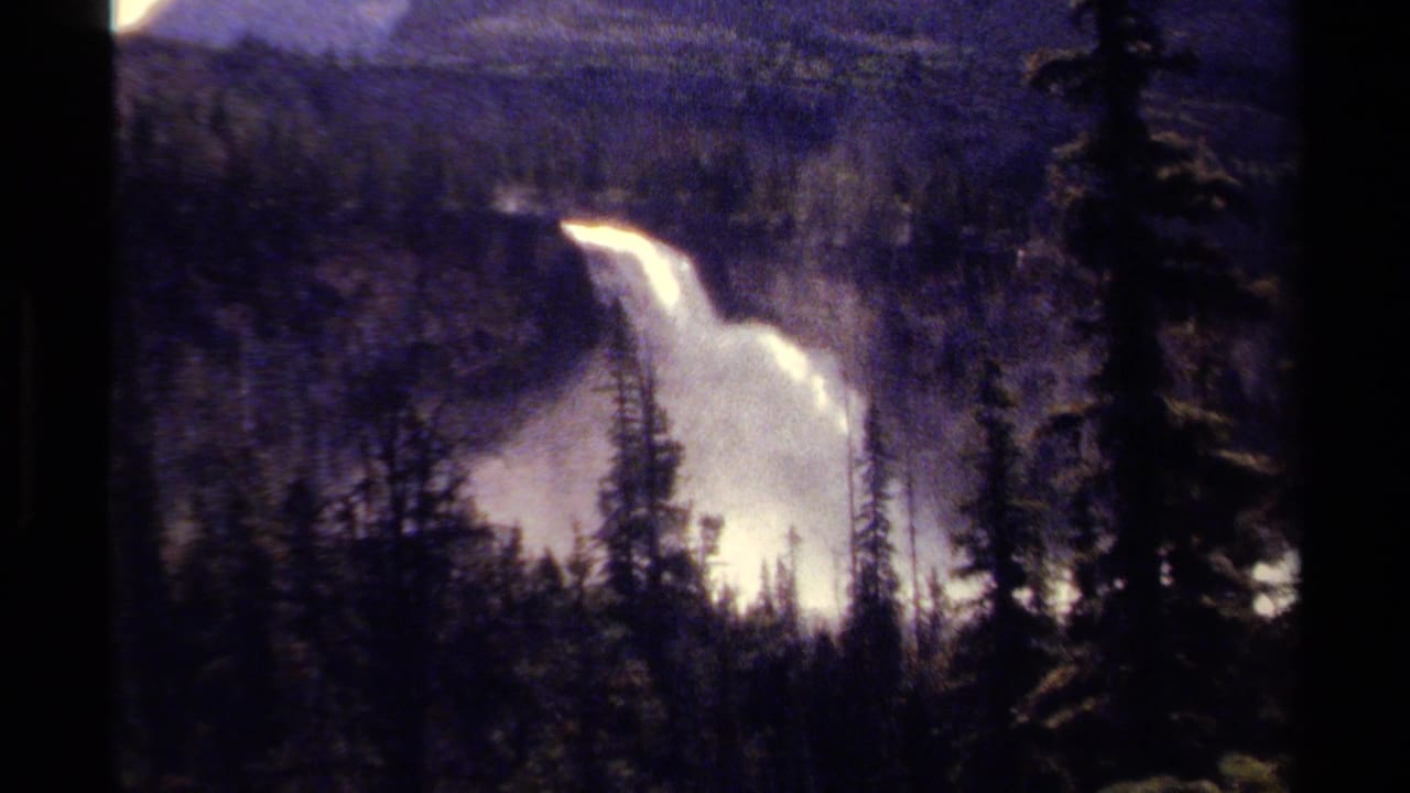 1979 8mm footage from British Columbia showing a distant waterfall framed by evergreens and hazy mountain slopes.