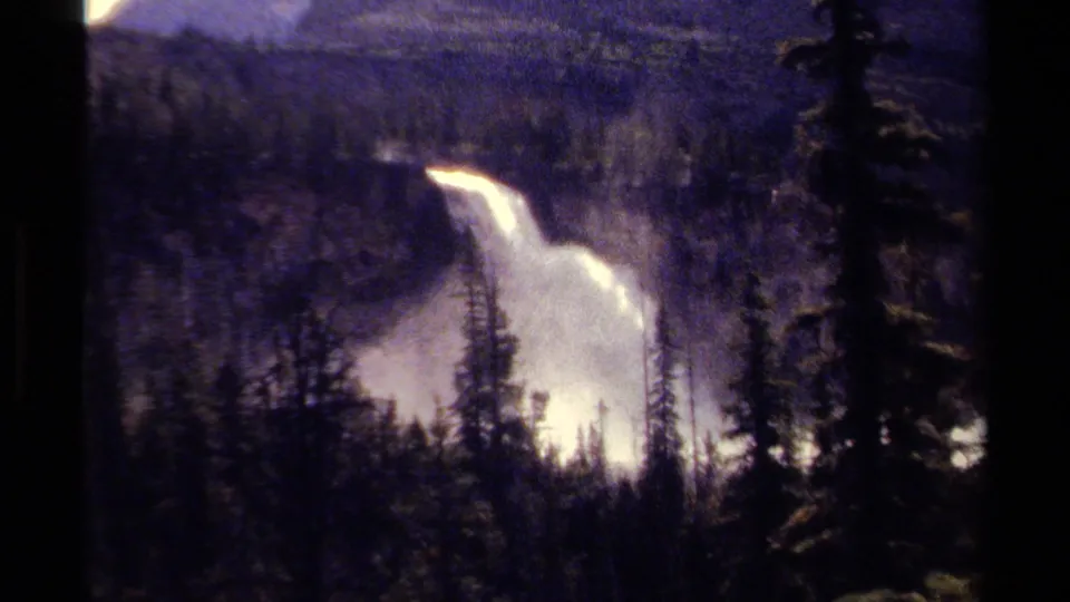 1979 8mm footage from British Columbia showing a distant waterfall framed by evergreens and hazy mountain slopes.