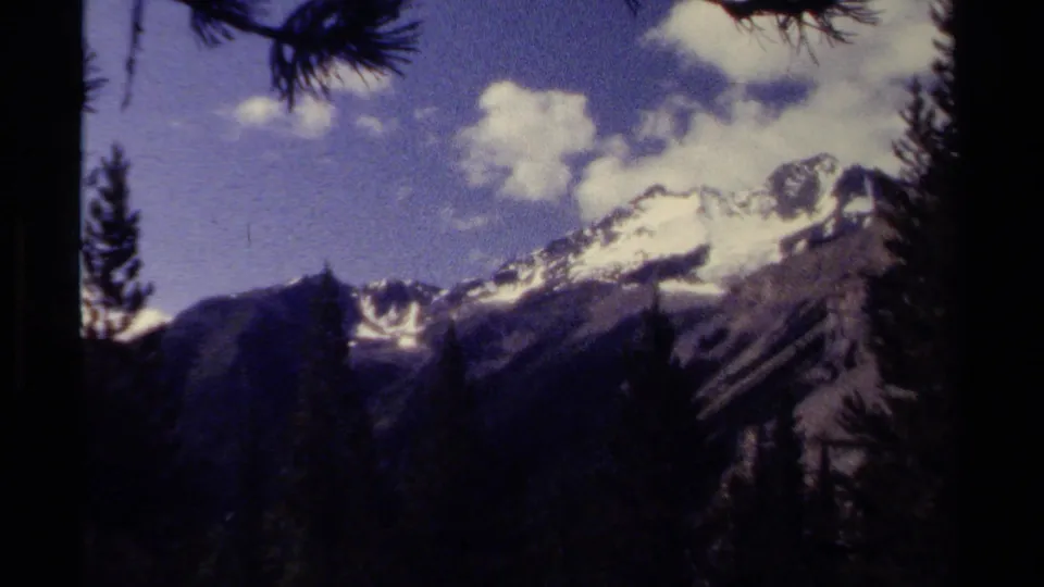 A British Columbia mountain peak appears through forest framing, with snow patches and clouds visible in the 8mm frame.