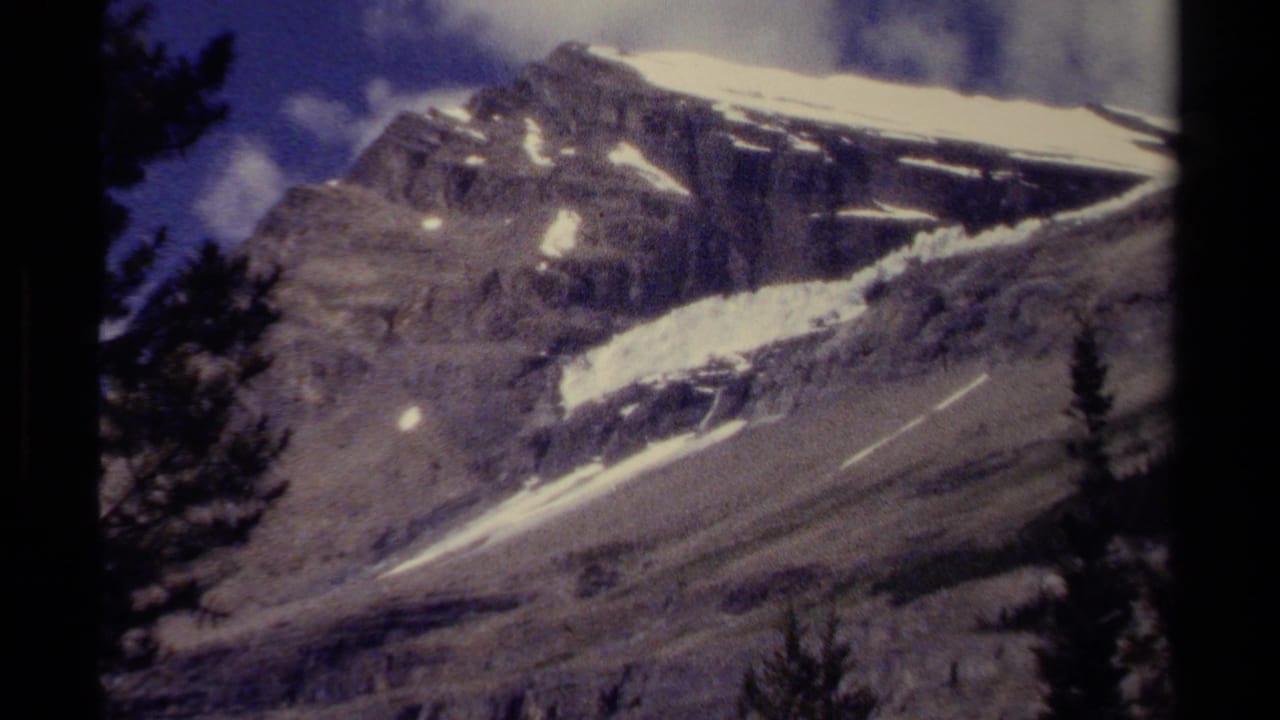 Snow-streaked British Columbia mountain peak under blue sky, framed by dark conifer shapes on 8mm film.