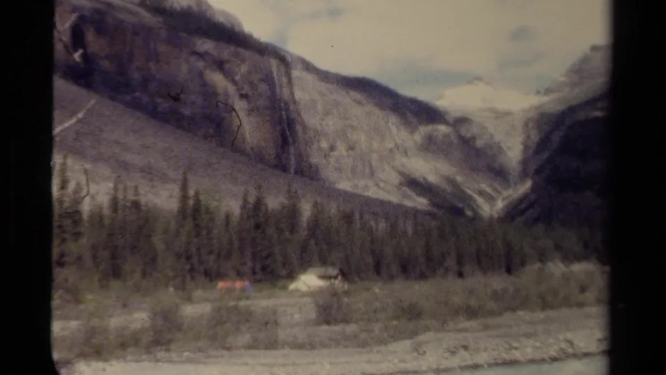 1979 British Columbia 8mm frame of a mountain valley with evergreen forest, riverbed, cliff face, waterfall, and small campsite.