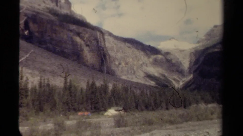 1979 British Columbia 8mm frame showing a broad mountain valley, forested slope, riverbed, cliff wall, and small campsite below.