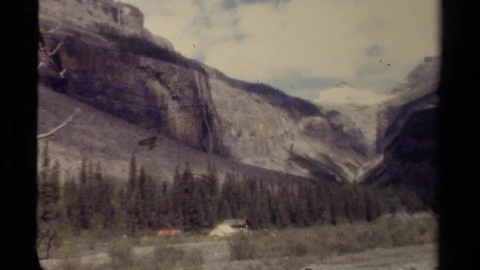 1979 British Columbia 8mm frame of a campsite at the base of cliffs, with evergreens, a riverbed, and waterfall in the mountain landscape.