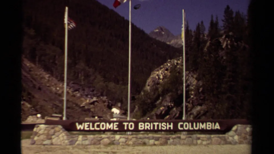 1979 8mm frame of a stone British Columbia welcome sign with flagpoles, forested slopes, and rocky mountain peaks.