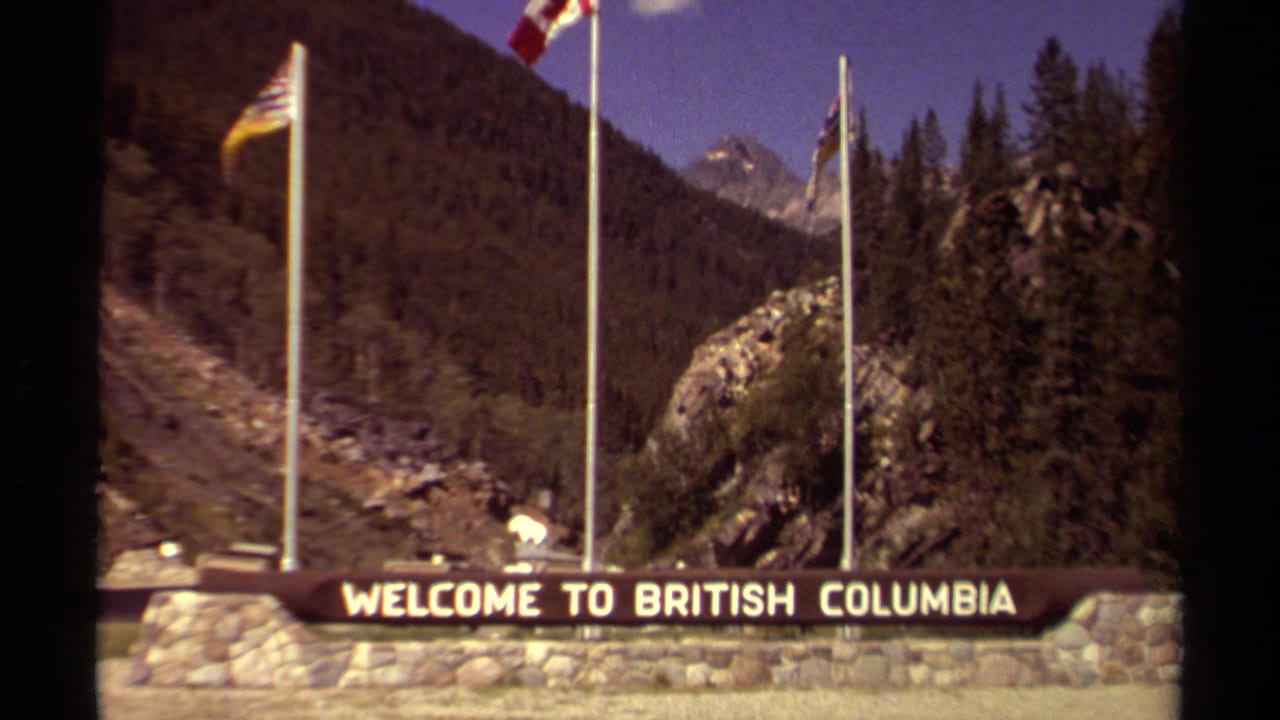 1979 British Columbia welcome sign on a stone base with flagpoles, evergreen slopes, and rocky mountain scenery behind it.