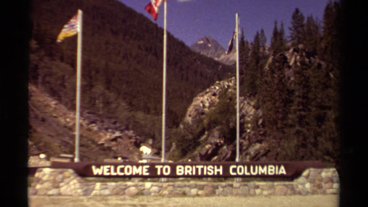 British Columbia welcome sign with flags, trees, rocks, and mountains in the background.