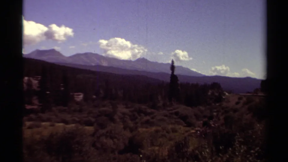 1979 British Columbia mountain scenery with dark evergreen forest, rolling foreground brush, distant peaks, and bright clouds.