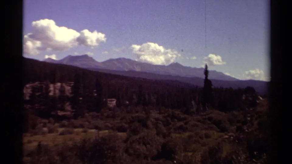 1979 British Columbia landscape showing a broad mountain range, evergreen trees, brushy terrain, and sunlit clouds in a blue sky.