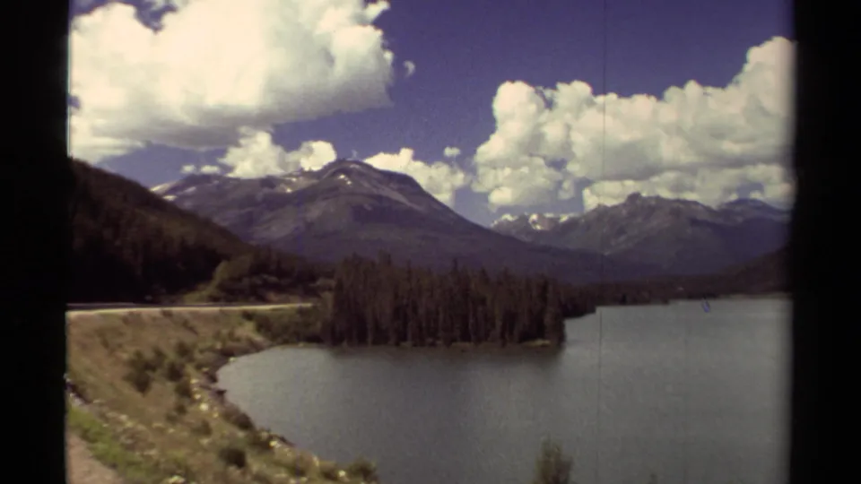 1979 British Columbia mountain lake scene with a curved shoreline road, evergreen trees, peaks, and large clouds.