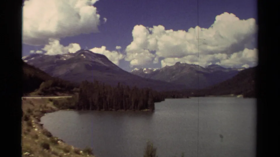 1979 British Columbia 8mm frame showing a calm lake, wooded shore, mountain range, and bright cumulus clouds.