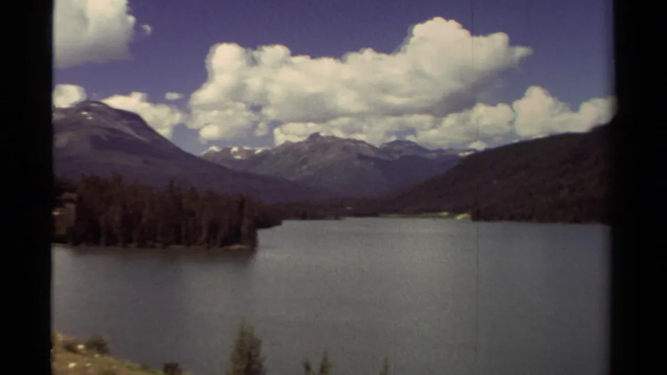 1979 British Columbia landscape with a broad lake, dark evergreen point, surrounding mountains, and cloud-filled sky.