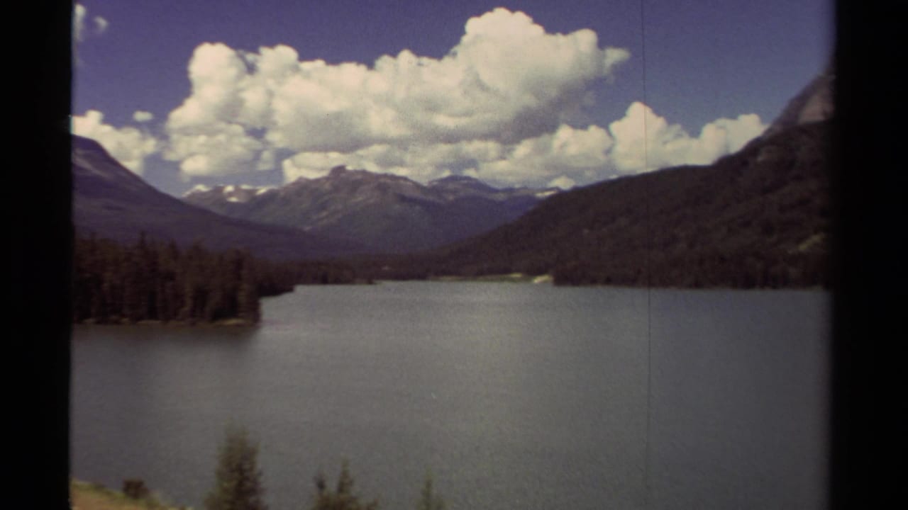 Mountain lake with evergreen shoreline, distant peaks, blue sky, and large white clouds.
