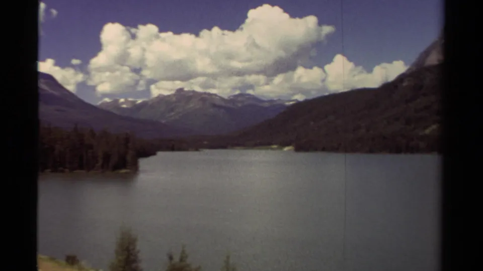 1979 8mm view of a British Columbia mountain lake with forested shores, distant peaks, and bright cumulus clouds.